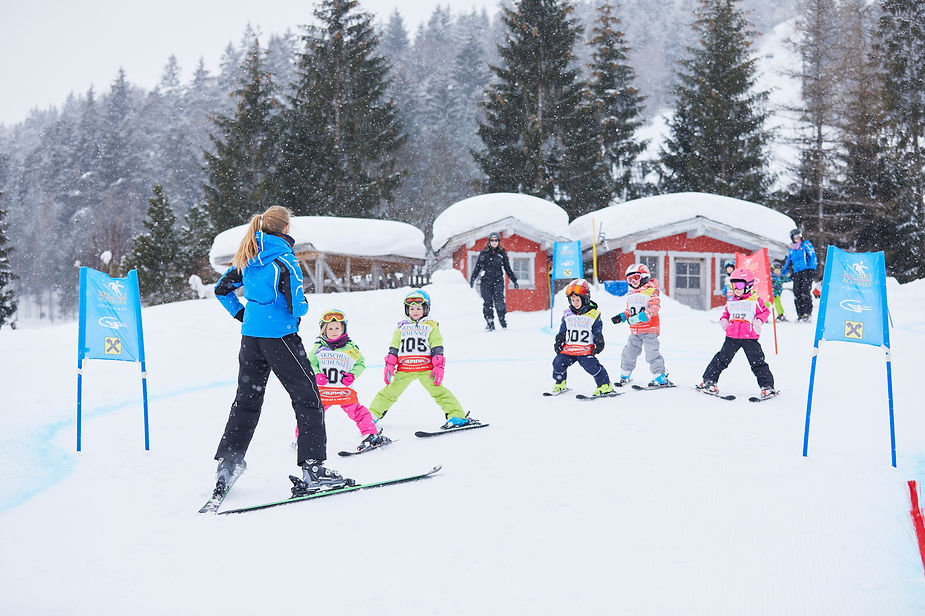 Skilehrerin mit blauer Jacke zeigt Kindern mit Skiern auf schneebedeckter Piste vor kleinen Hütten eine Übung