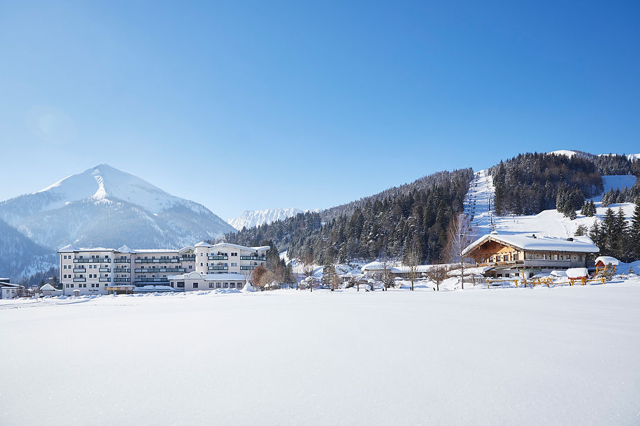 Schneebedeckte Landschaft mit mehreren Gebäuden vor bewaldeten Bergen und klarem Himmel