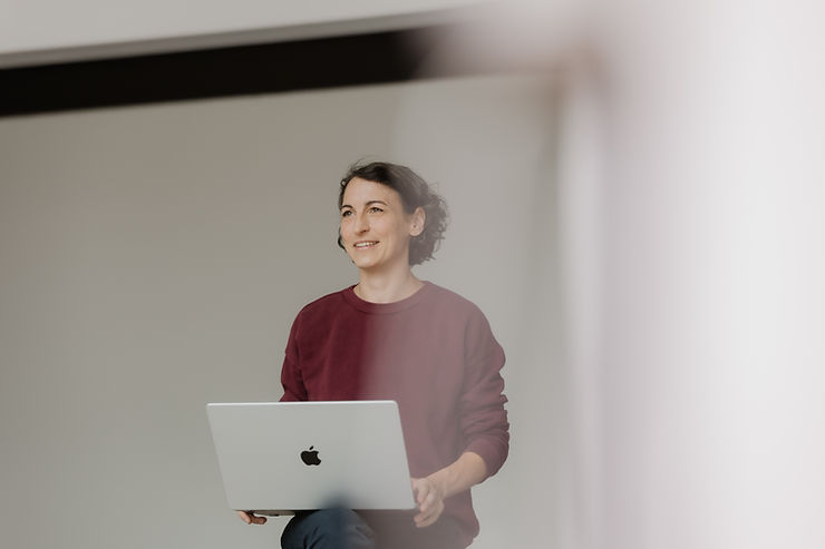 Natascha Wegelin mit lockigem Haar und rotem Pullover sitzt mit einem aufgeklappten Apple-Laptop auf dem Schoß