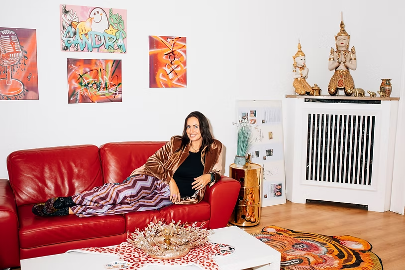 Rechtsanwältin Sandra Günther liegt entspannt auf einem roten Ledersofa in einem Raum mit bunten Wandbildern, einem weißen Tisch mit Dekoration und einer goldfarbenen Skulptur auf einem Heizkörper.