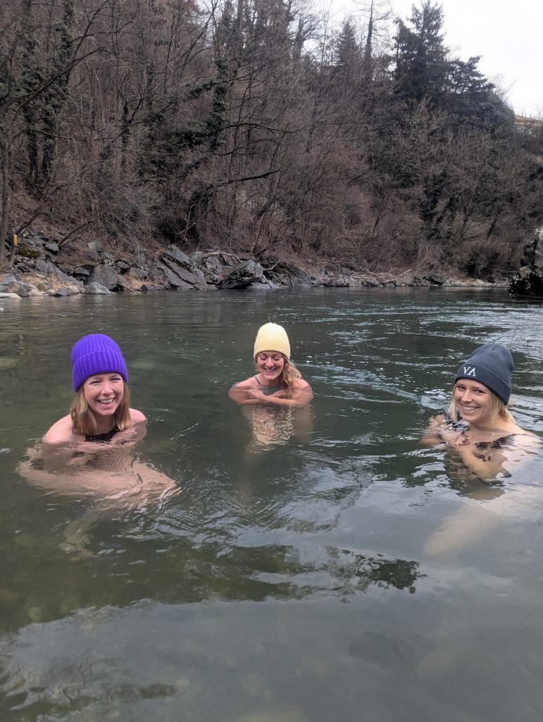 Drei Frauen mit Mützen sitzen im Flusswasser vor bewaldetem Ufer