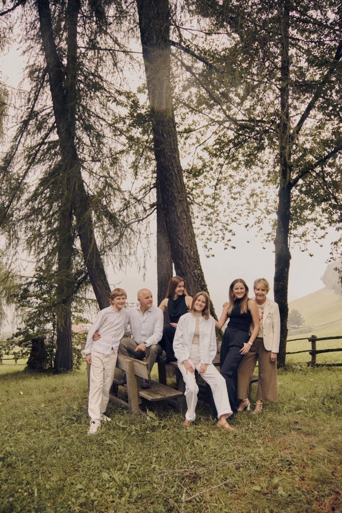 Sechs Personen unterschiedlicher Altersgruppen stehen und sitzen auf einer Holzbank vor mehreren hohen Bäumen auf einer Wiese mit Holzzaun im Hintergrund
