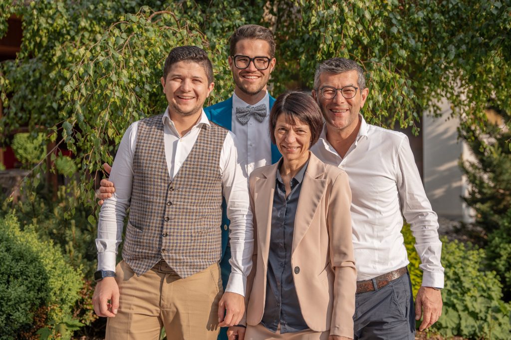 Vier Personen stehen vor einem Baum mit hängenden Zweigen, drei Männer tragen Hemden, einer davon mit Weste und einer mit Fliege, eine Frau trägt einen Blazer über einer Bluse