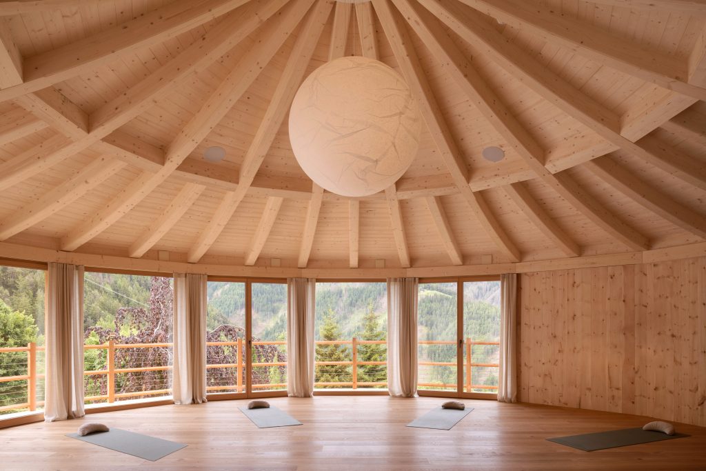 Runder heller Raum mit Holzwänden und sichtbaren Dachbalken, großen Fenstern mit Vorhängen und Yogamatten auf dem Boden