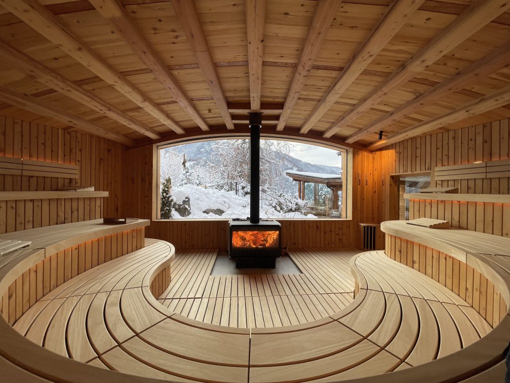 Saunaraum mit halbkreisförmigen Holzbänken, Holzofen mit sichtbarem Feuer und großem Fenster mit Blick auf verschneite Landschaft