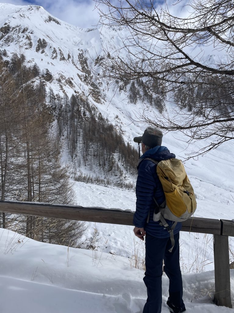 Wanderer mit gelbem Rucksack und blauer Jacke steht vor schneebedecktem Berg und blickt nach oben