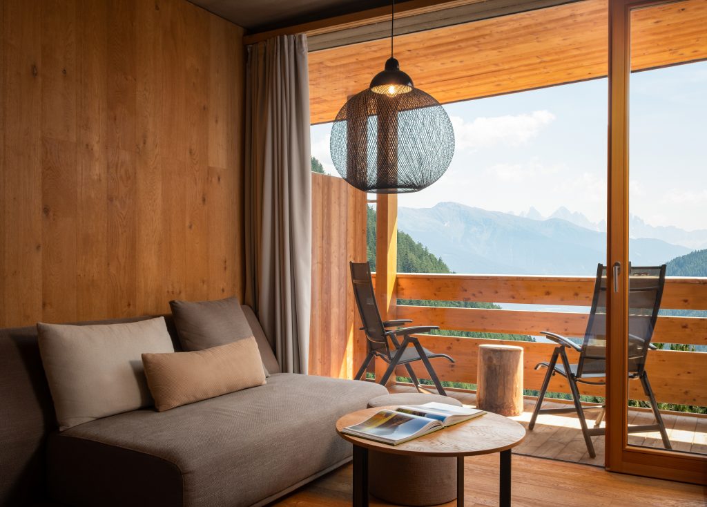 Wohnzimmer mit brauner Couch, rundem Holztisch, zwei Stühlen auf dem Balkon und Bergblick durch große Glas-Schiebetür