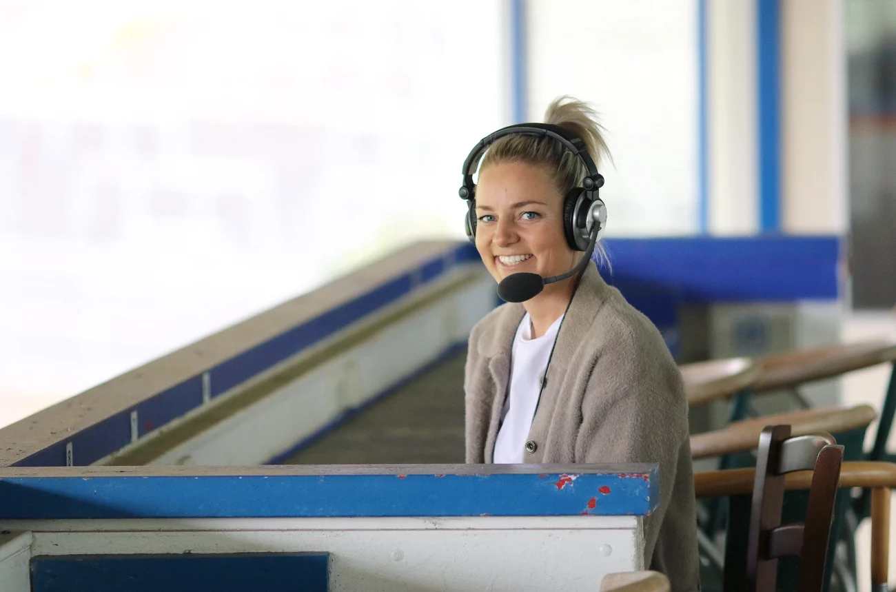 Christina Graf mit Kopfhörern sitzt an einem blauen Holztisch in einem hellen Raum mit großen Fenstern