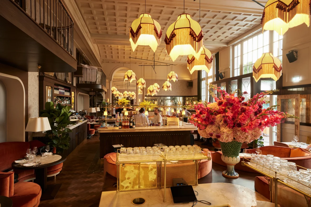 Großer, eleganter Gastraum mit hohen Fenstern, mehreren hängenden Lampen, einer Bar in der Mitte und einem großen Blumenarrangement auf einem Tisch