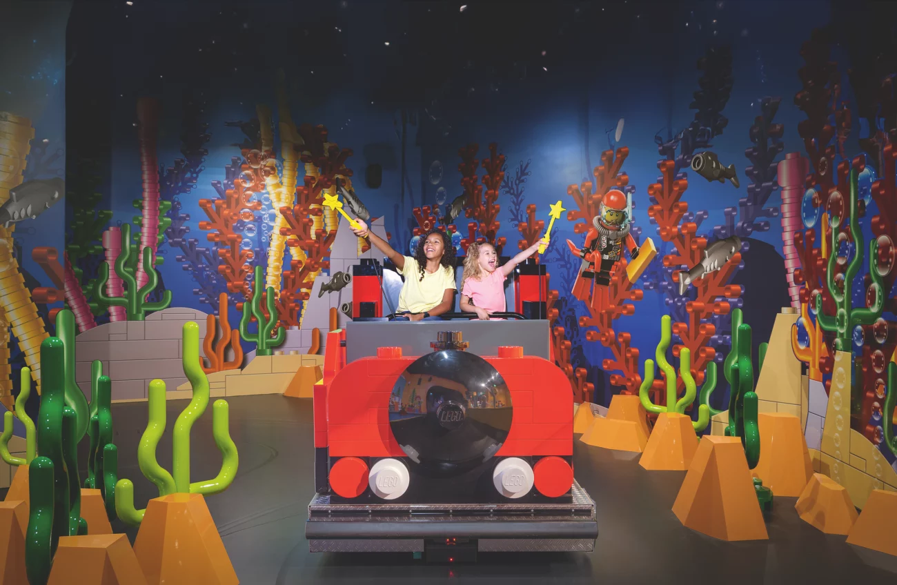 Zwei Personen sitzen in einem roten Lego-U-Boot-Modell vor einer bunten Unterwasser-Lego-Kulisse mit Korallen und Fischen.