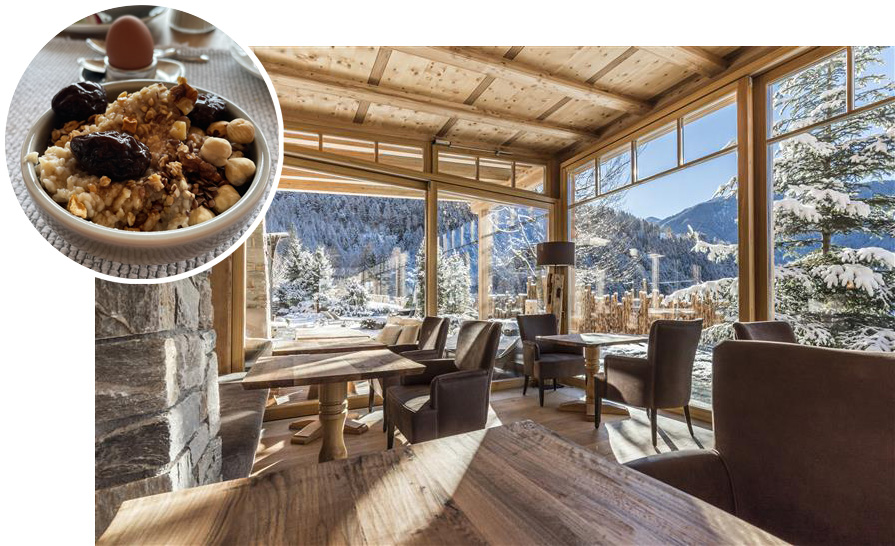 Gemütlicher Frühstücksraum mit Holzmöbeln und großen Fenstern, die auf verschneite Berge und Tannen blicken, dazu eine Schale mit Haferbrei, Nüssen und Trockenfrüchten sowie ein gekochtes Ei im Hintergrund