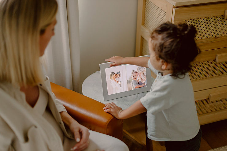 Kind steht an einem kleinen runden Tisch und zeigt auf einen digitalen Bilderrahmen mit Familienfotos