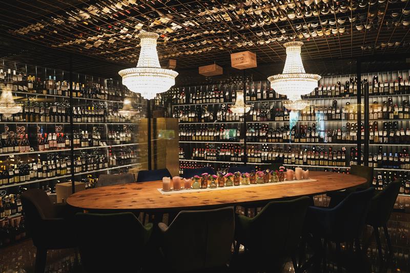 Großer ovaler Holztisch mit mehreren Stühlen in einem Raum, dessen Wände mit Regalen voller Weinflaschen bedeckt sind und von mehreren Kronleuchtern beleuchtet werden