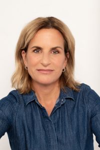 Claudia Kettler mit schulterlangem blonden Haar trägt ein blaues Jeanshemd und silberne Ohrringe vor hellem Hintergrund