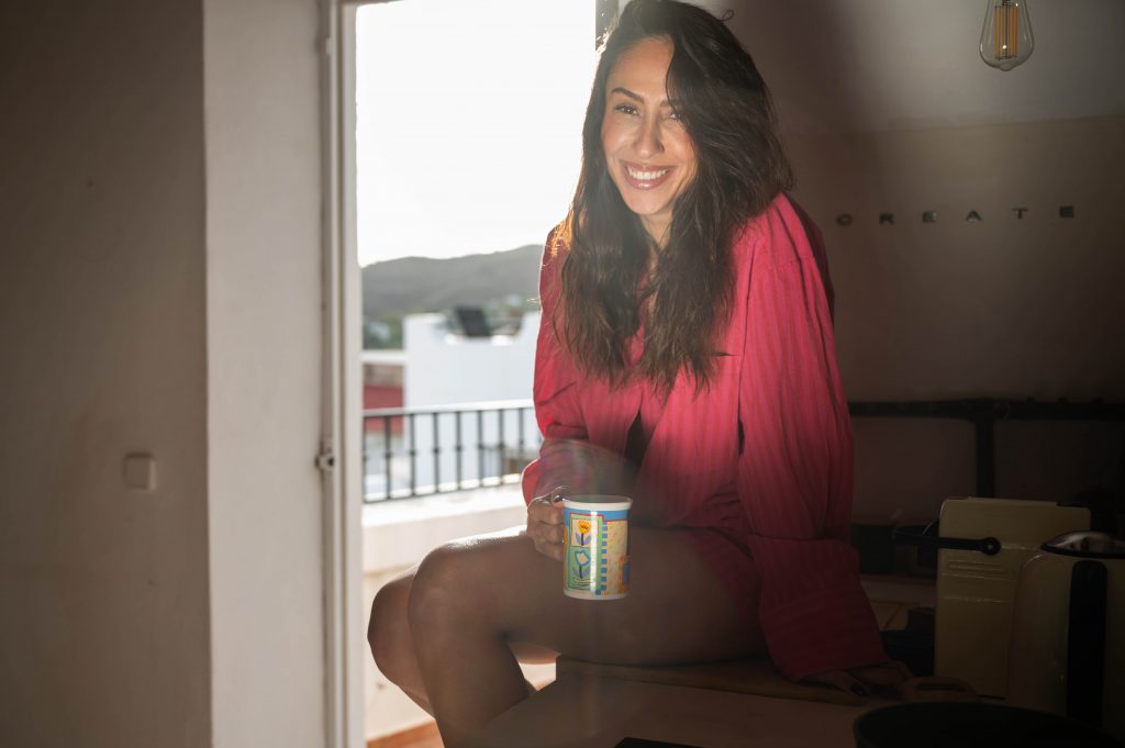 Melissa Khalaj mit langem Haar und rotem Oberteil sitzt auf einer Fensterbank und hält eine bunte Tasse