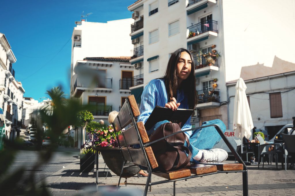 Melissa Khalaj sitzt auf einer Bank in urbaner Umgebung und schreibt in ein Notizbuch