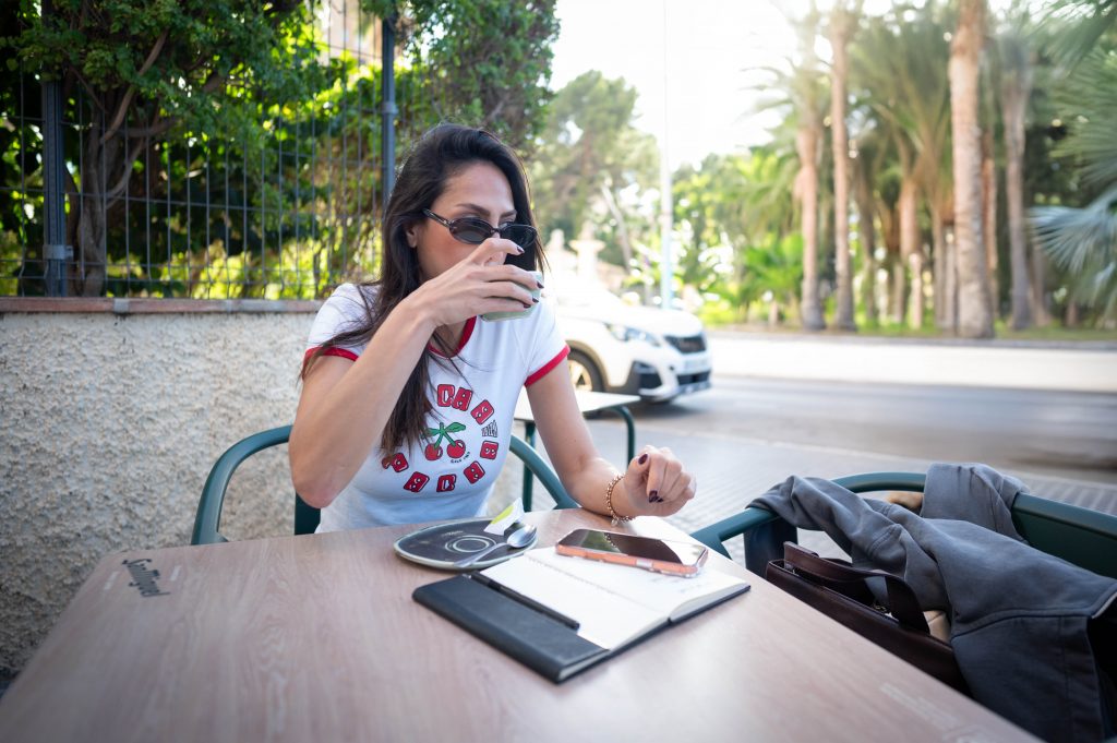 Melissa Khalaj sitzt an einem Tisch im Freien mit Smartphone, Notizbuch und Stift vor sich und hält sich die Hand vor den Mund