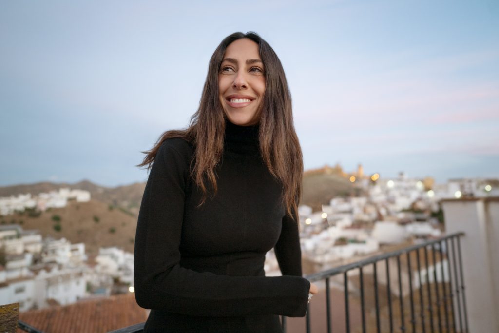 Melissa Khalaj mit langem Haar und schwarzem Rollkragenpullover lehnt an einem Geländer vor einer Stadtlandschaft mit weißen Häusern auf Hügeln