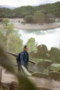 Melissa Khalaj mit Rucksack und Jeansjacke steht auf einem Pfad mit Geländer vor einem See und bewaldeten Hügeln im Hintergrund