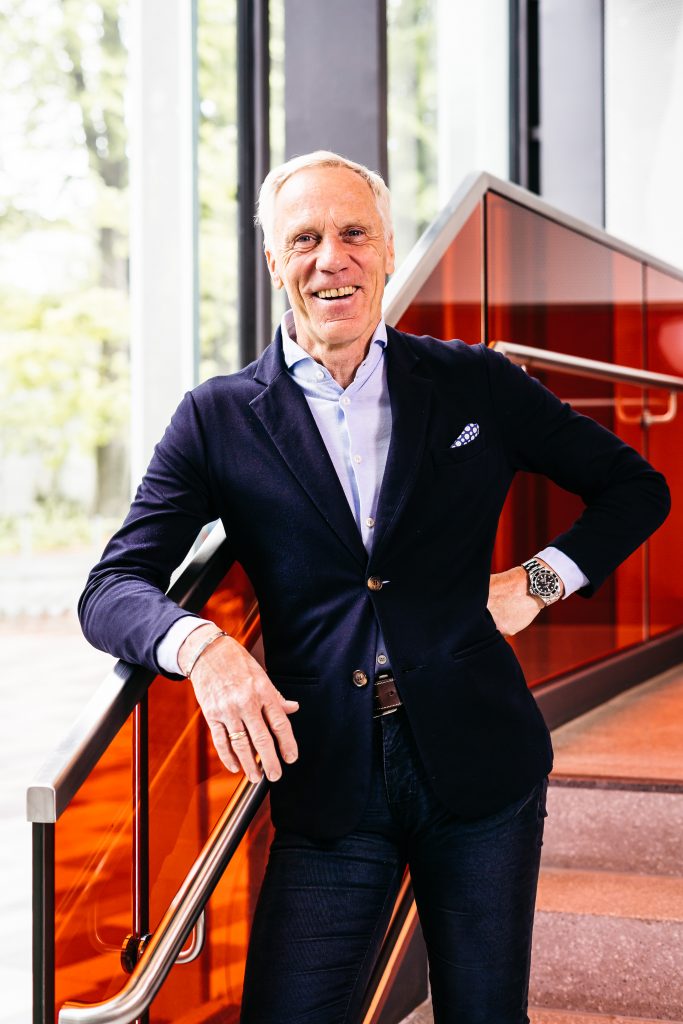 Ingo Froböse in dunklem Sakko lehnt an Treppengeländer mit orangefarbenen Glasfüllungen in modernem Gebäude