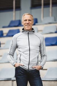 Ingo Froböse steht mit Händen in den Hosentaschen vor Reihen von Sitzplätzen in einer Sportarena