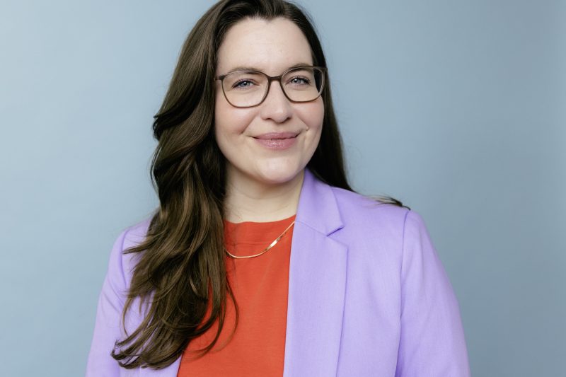 Madita Oeming mit langen braunen Haaren trägt ein helles Sakko und ein rotes Oberteil vor hellem Hintergrund