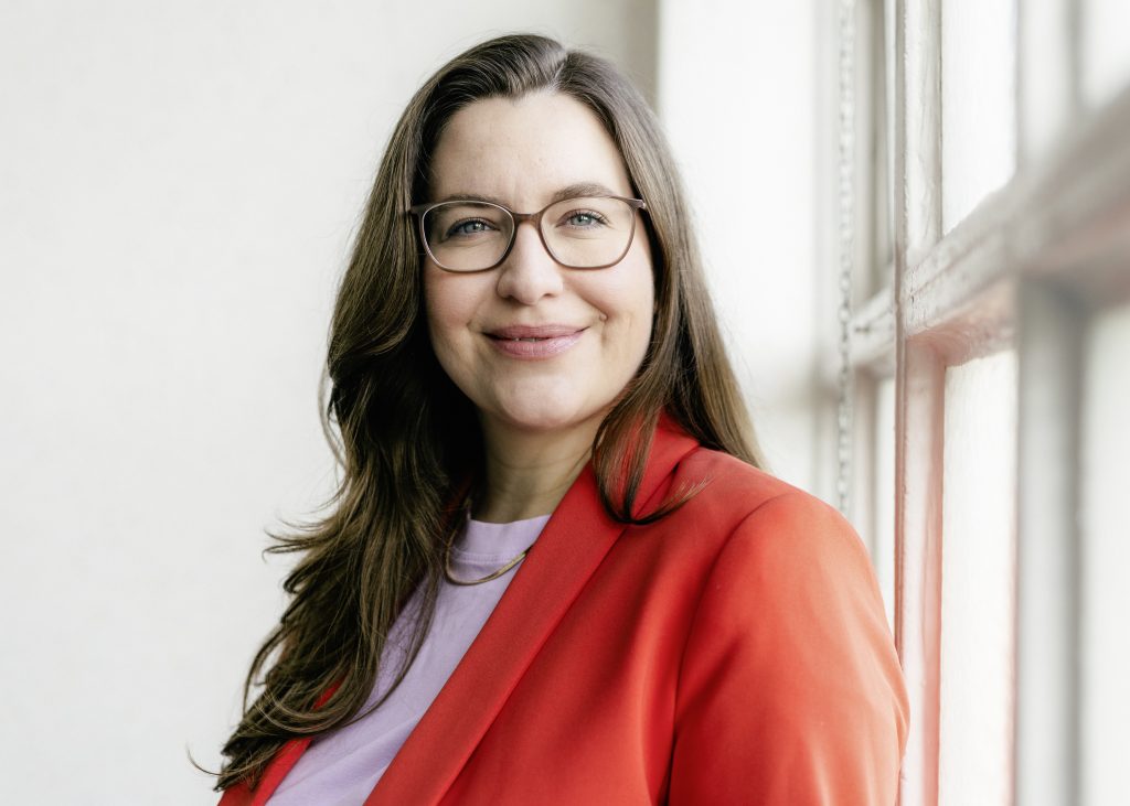 Madita Oeming mit langen braunen Haaren trägt roten Blazer und helles Oberteil steht seitlich an einem Fenster