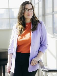 Madita Oeming mit schulterlangem Haar trägt orangefarbenes Oberteil und hellen Blazer lehnt an Geländer in hellem Raum