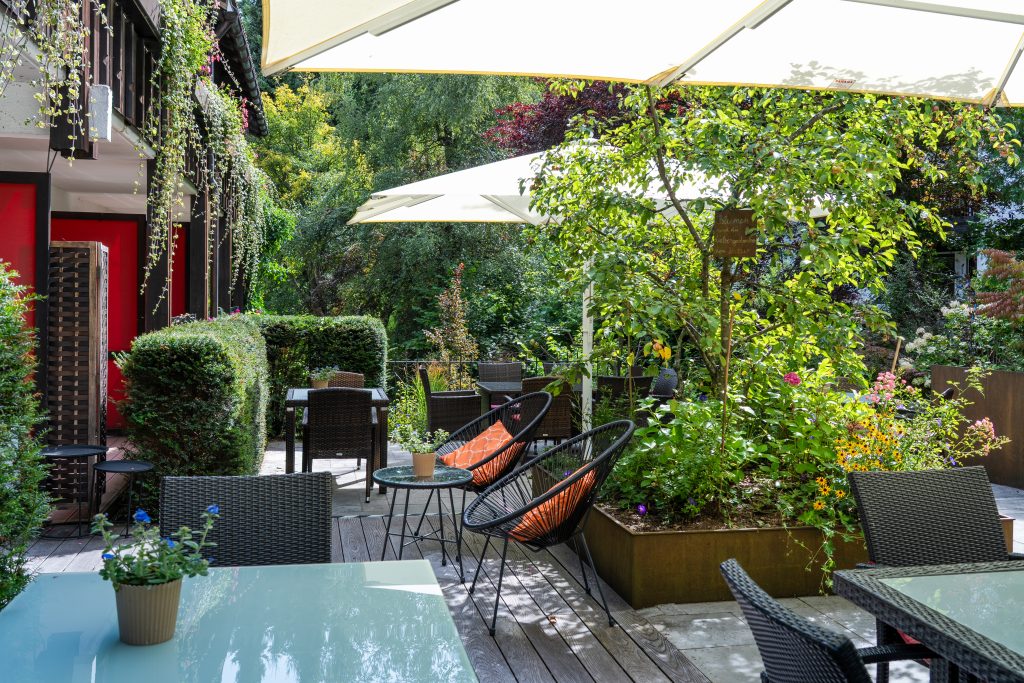 Terrasse mit Tischen, Stühlen und Sonnenschirmen umgeben von grünen Pflanzen und Bäumen