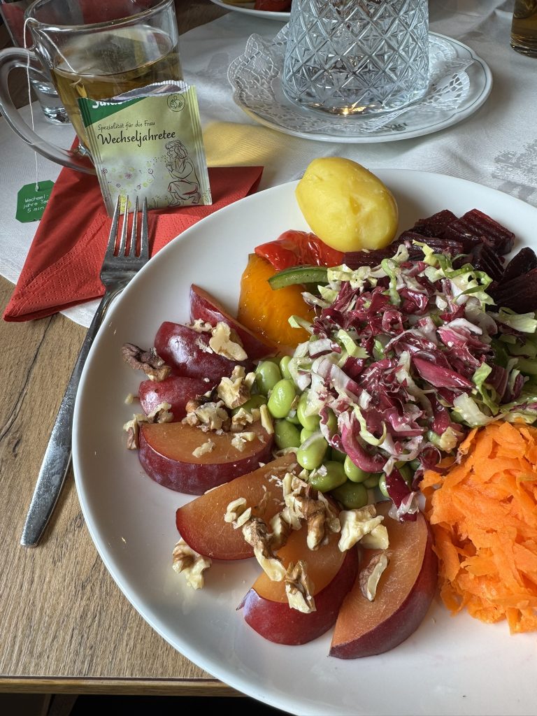 Teller mit Salat aus Radicchio, Karotten, Roter Bete, Edamame, Pflaumenstücken mit Walnüssen und einem kleinen Kartoffelknödel auf Holztisch mit Besteck und Teetasse
