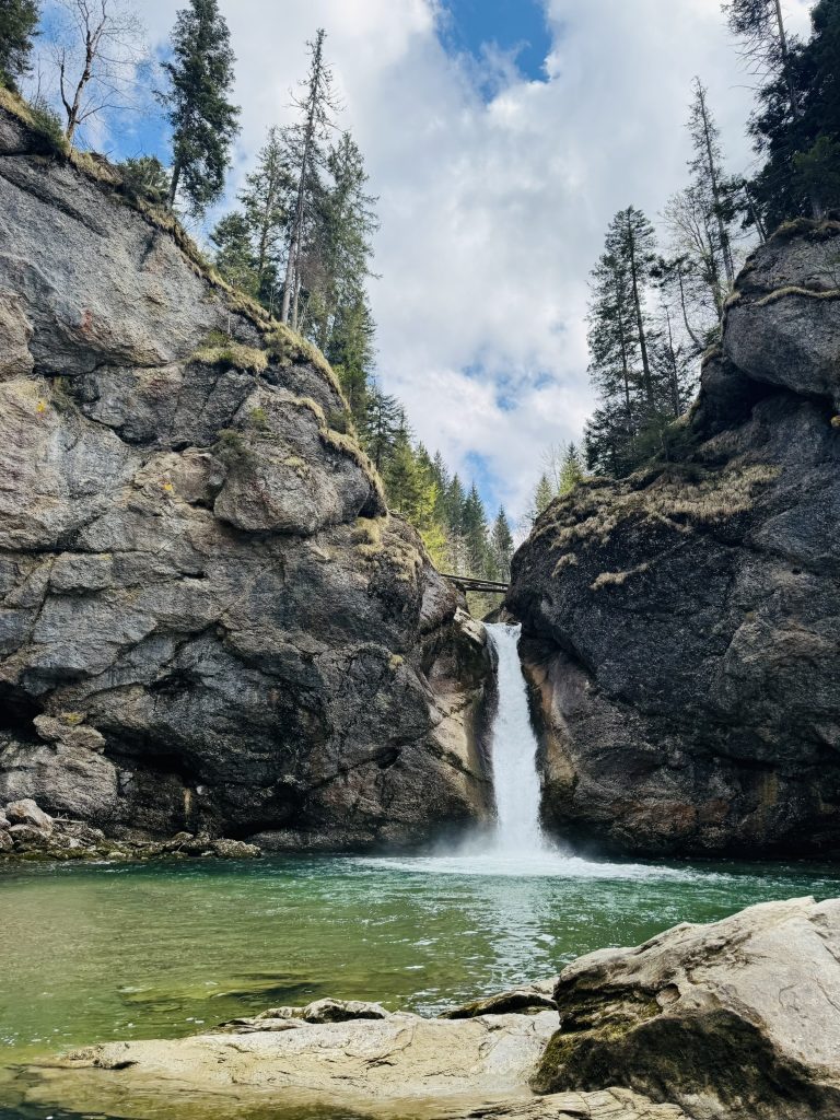 Wasserfall zwischen zwei großen Felsen mit grünem Wasser und Bäumen auf den Felsen unter bewölktem Himmel