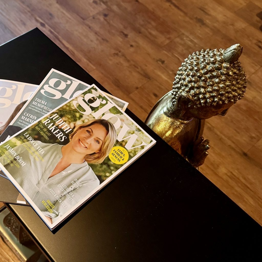 Goldfarbene Buddha-Statue neben einem schwarzen Tisch mit mehreren Ausgaben der Zeitschrift "glow" auf einem Holzboden
