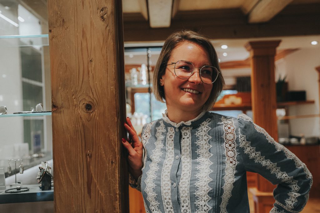 Sandra Ammer lehnt mit einer Hand an einem Holzpfosten in einem Raum mit Regalen und Säulen und trägt eine gemusterte Bluse mit Spitzenbesatz