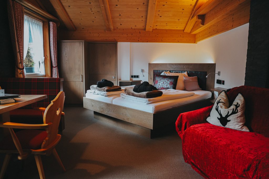 Gemütliches Schlafzimmer mit Holzbalkendecke, Doppelbett, rotem Sessel mit Kissen und Holzmöbeln