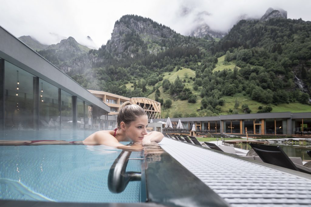 Frau lehnt entspannt an der Kante eines Infinity-Pools mit Berglandschaft im Hintergrund