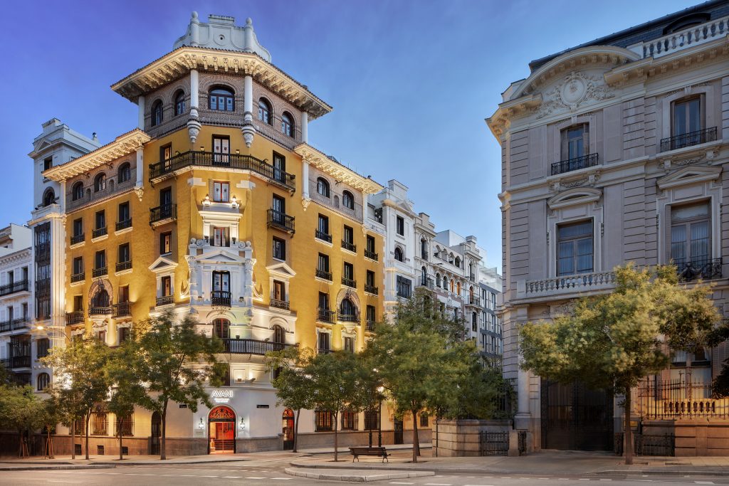 Mehrstöckiges historisches Gebäude mit gelber Fassade an einer Straßenecke, umgeben von Bäumen und weiteren Gebäuden