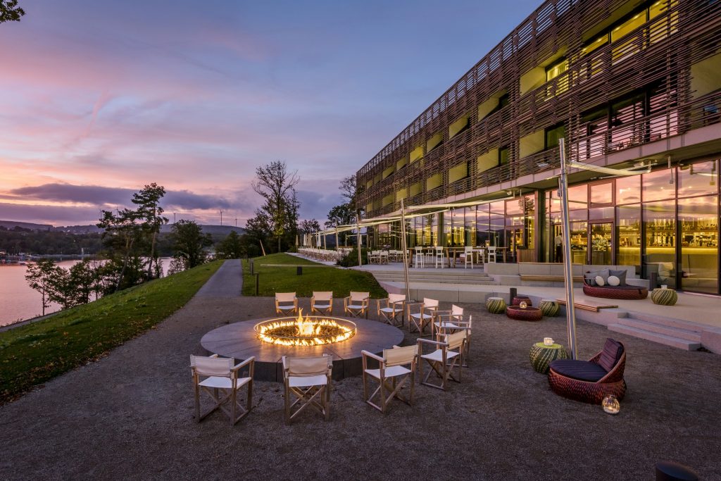 Feuerstelle mit brennendem Feuer und Stühlen im Kreis vor einem modernen Hotelgebäude am Seeufer bei Sonnenuntergang