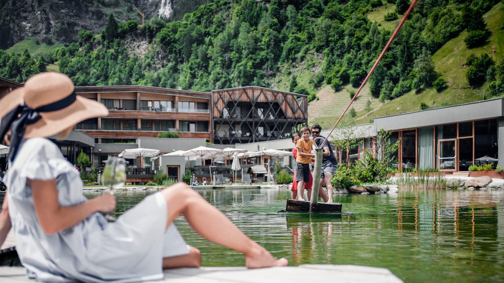 Frau mit Hut und hellblauem Kleid sitzt am Ufer eines Sees, im Hintergrund zwei Personen auf einer Seilfähre über das Wasser