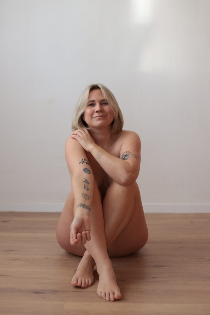 Nackte sitzende Louisa Dellert mit blonden Haaren und mehreren sichtbaren Tätowierungen auf Armen und Beinen vor weißer Wand und Holzboden