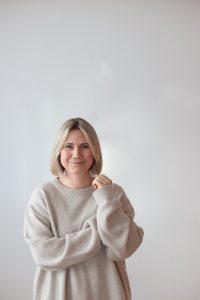 Louisa Dellert mit schulterlangem blonden Haar trägt einen hellen, grob gestrickten Pullover und hält eine Hand nahe am Kinn