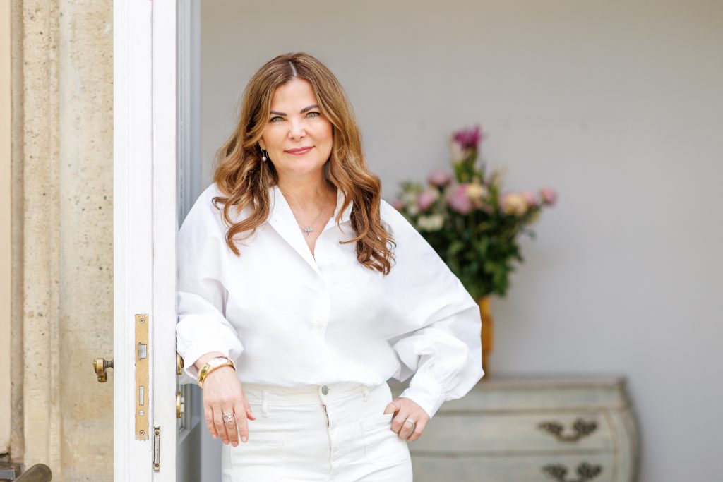 Dr. med. Sheila de Liz mit langen, gewellten Haaren in weißer Bluse und weißer Hose lehnt an einer Tür, im Hintergrund eine Kommode mit Blumenstrauß