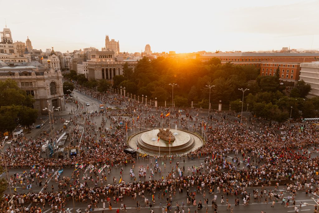 Luftaufnahme eines großen Platzes mit einem runden Brunnen in der Mitte und vielen Menschen, die sich um den Brunnen und auf den umliegenden Straßen versammeln, bei Sonnenuntergang