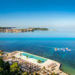 Infinity-Pool mit Liegen und Sonnenschirmen an der Küste mit Blick auf das Meer und eine entfernte Stadt