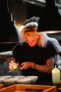 Ana Roš mit blonden Haaren reibt Zitrusfrucht über eine Reibe vor mehreren weißen Schalen in einer Küche