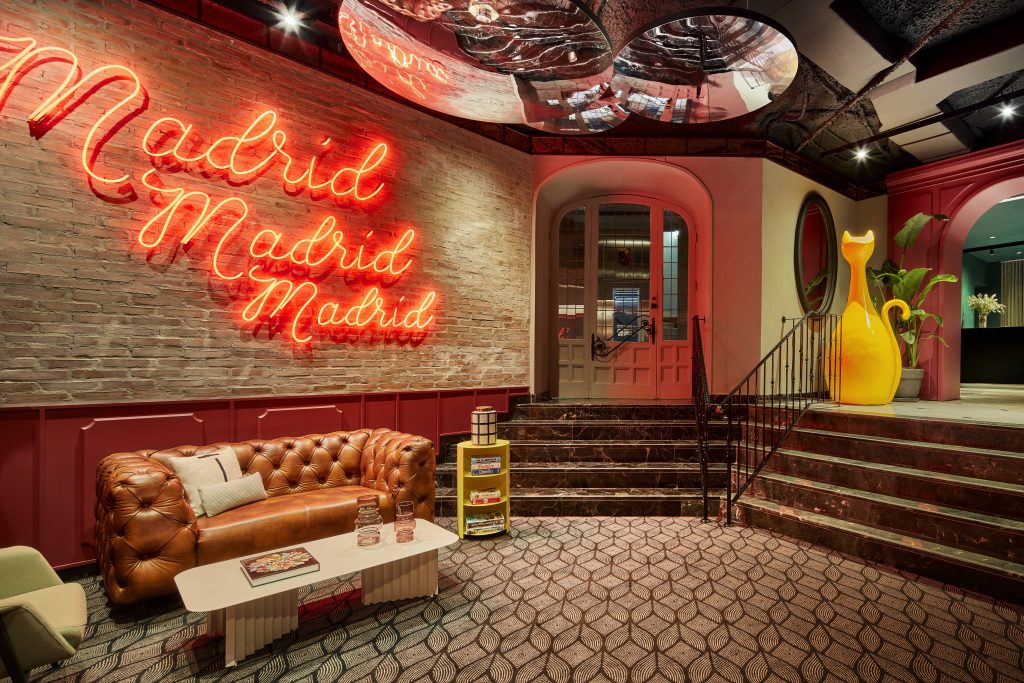 Lounge mit braunem Ledersofa, weißem Tisch, roter Neonbeleuchtung mit Schriftzug 'Madrid' an Ziegelwand und gelber Skulptur neben Treppe