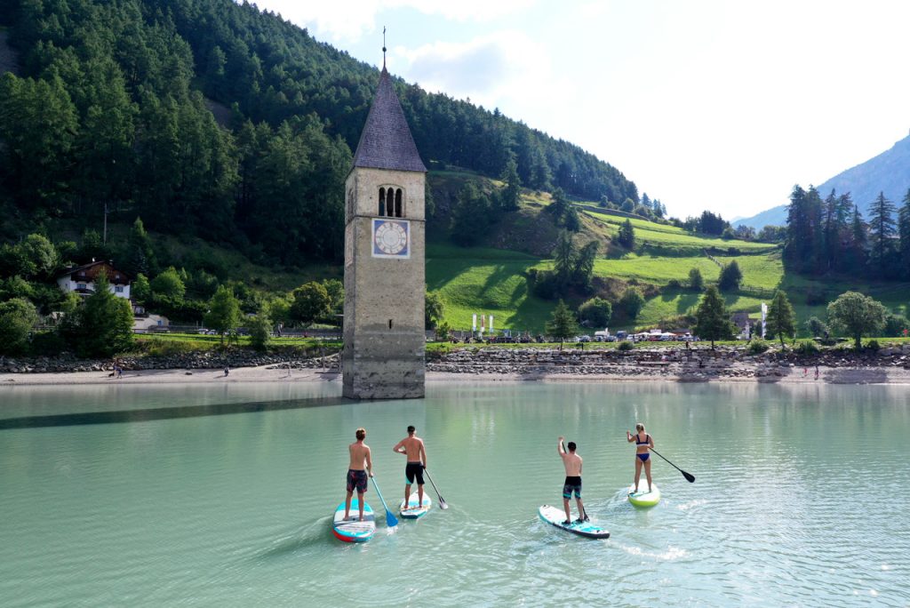 Vier Personen auf Stand-up-Paddle-Boards auf einem See vor einem teilweise unter Wasser stehenden Kirchturm in einem Bergtal