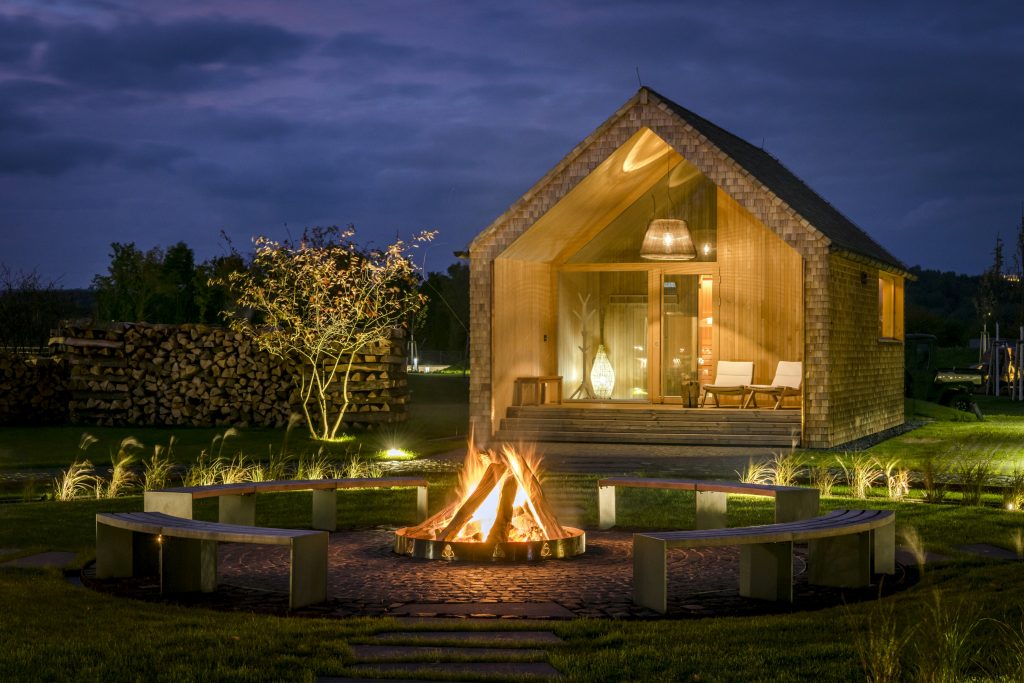 Kleines Holzhaus mit beleuchtetem Innenraum und davor ein brennendes Lagerfeuer bei Nacht