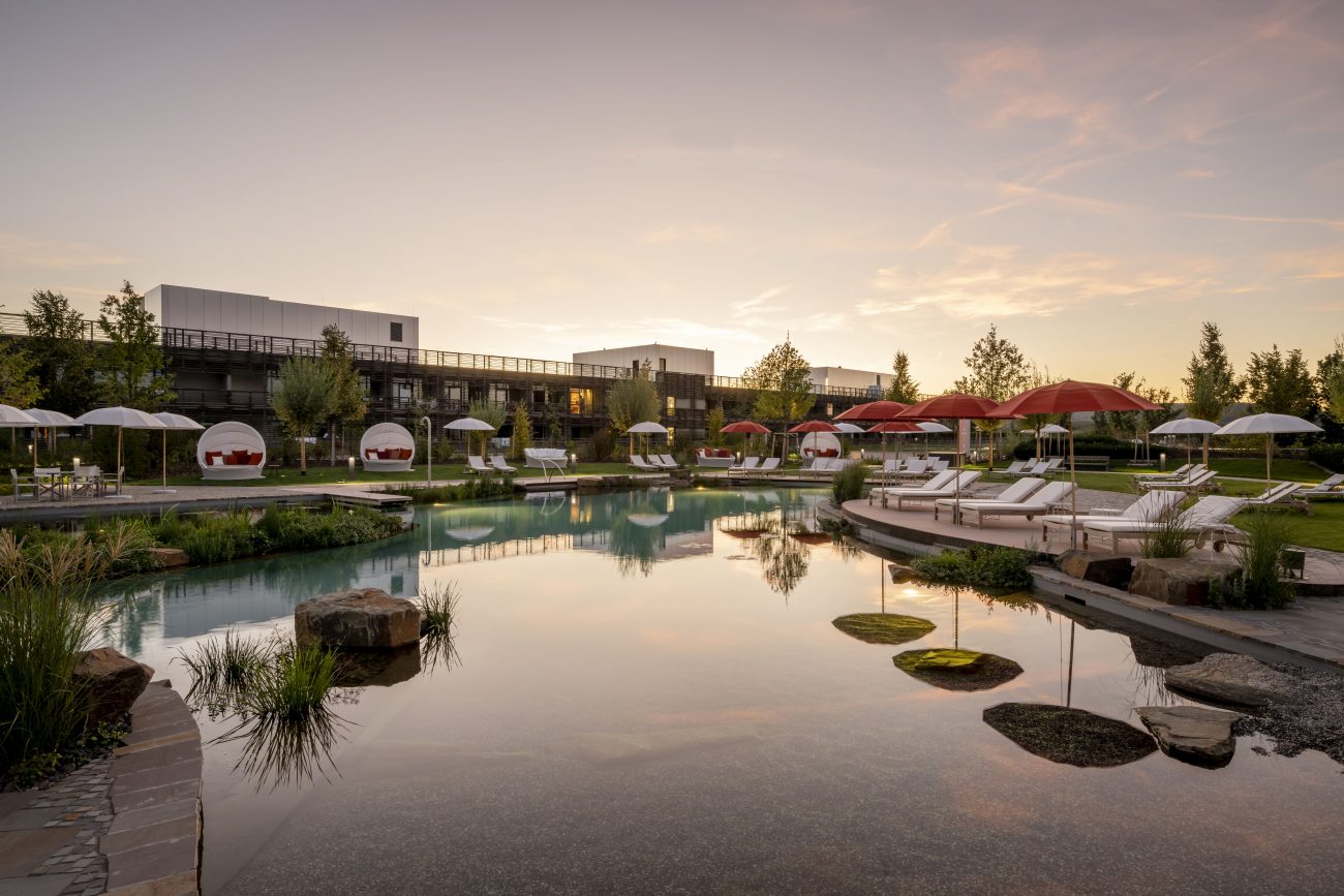 Naturnaher Badeteich mit Liegen und Sonnenschirmen vor modernem Hotelgebäude bei Sonnenuntergang