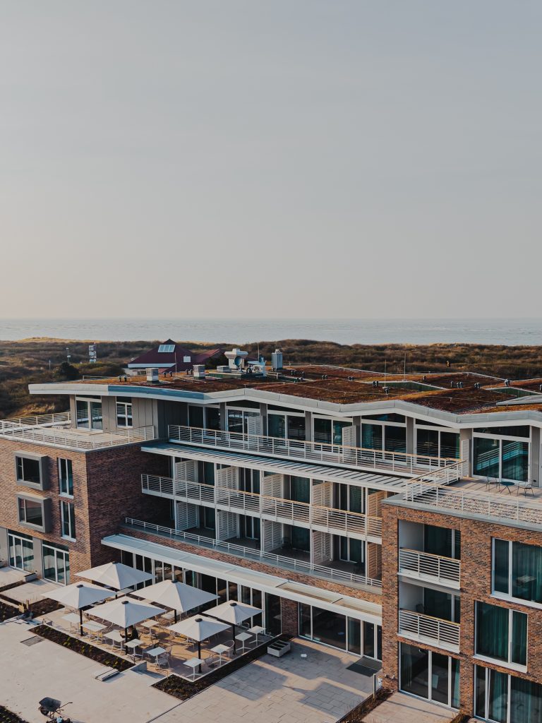 Mehrstöckiges Gebäude mit begrüntem Flachdach und mehreren Balkonen vor einer Dünenlandschaft