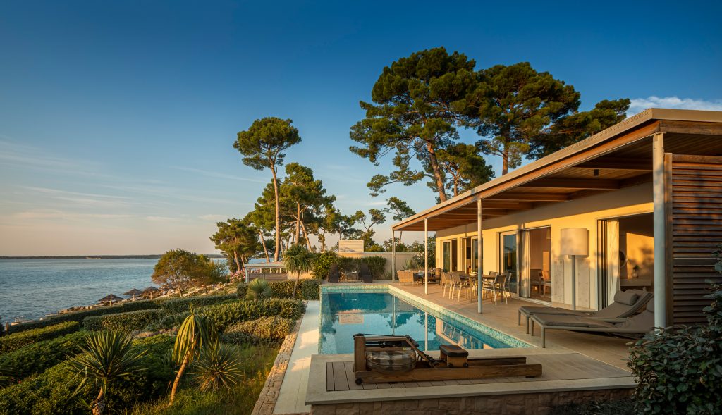 Moderner Bungalow mit Terrasse, Pool und Liegestühlen vor Meer und Bäumen bei Sonnenuntergang